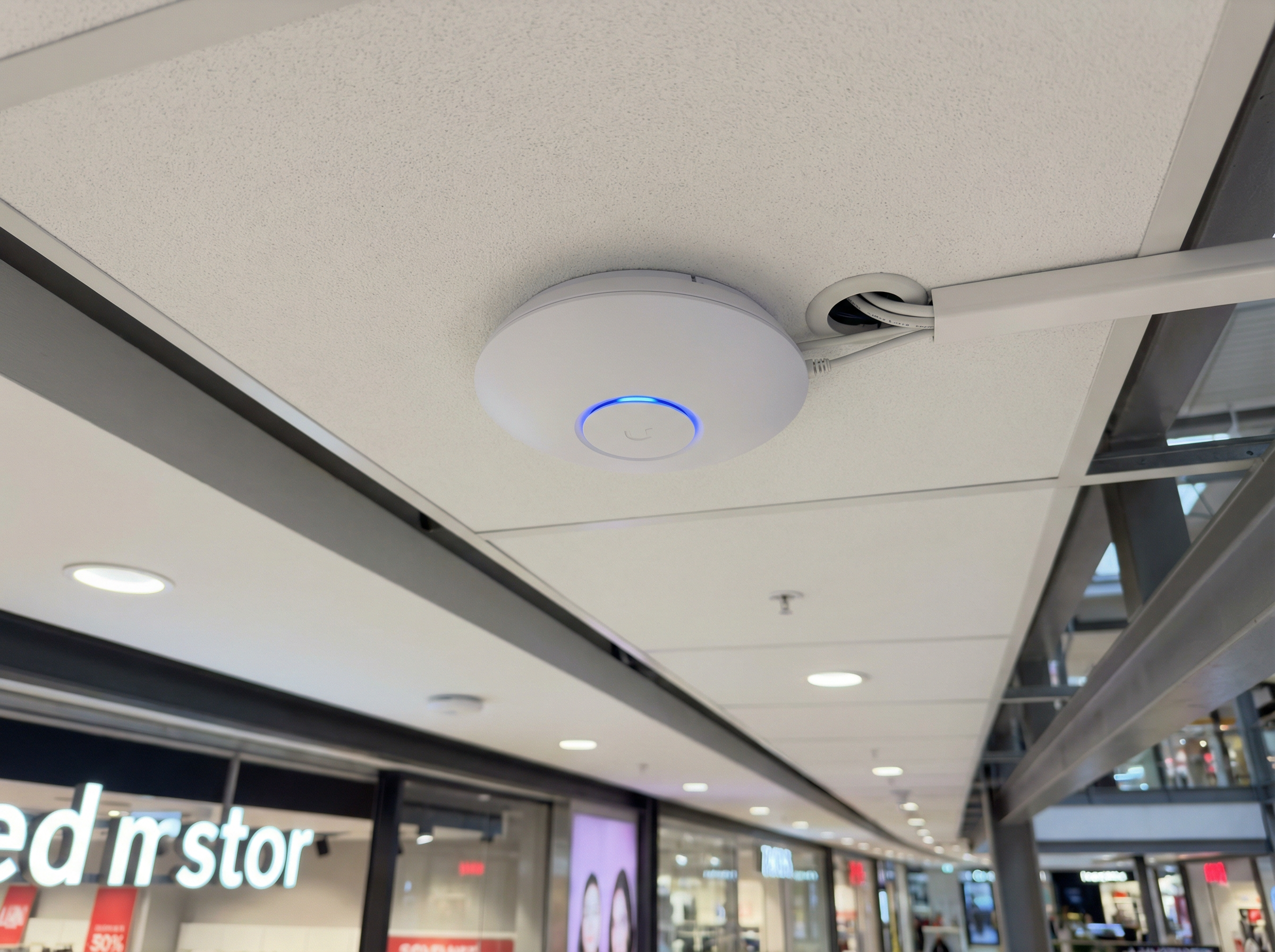 Access point Wi-Fi 6E enterprise instalado no teto de um shopping center em São Paulo, com LED indicador azul e gerenciamento de cabos profissional